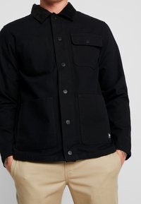 Schwarze Jacke aus Baumwolle mit Cordkragen, ausgestattet mit zwei oberen Taschen und zwei unteren Taschen, Knopfverschluss und einer strukturierten Form.