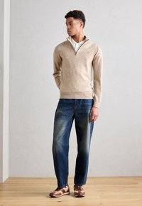 Pullover lavorato a maglia beige con colletto a mezza zip, abbinato a jeans blu a gamba dritta e sneakers marroni con dettagli a righe e lacci blu.