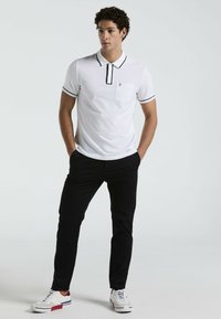 Weißes Polo-Shirt mit schwarzem Besatz und Tasche, kombiniert mit schwarzen Hosen und weißen Sneakers. Lässiges Design mit kurzen Ärmeln und taillierter Form.