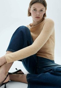 Beige knitted sweater with a round neckline, paired with dark blue denim jeans, featuring a subtle faded texture and a relaxed silhouette.