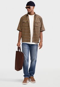 Πουκάμισο με κοντά μανίκια και καρό μοτίβο σε καφέ και κίτρινο, λευκό T-shirt, σκουρόχρωμο μπλε τζιν, καφέ τσάντα tote και καφέ αθλητικά παπούτσια με λευκές ρίγες.