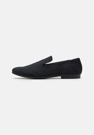 Mocassin à enfiler en daim noir avec un talon bas et un bout légèrement pointu, présenté de profil sur un fond blanc.