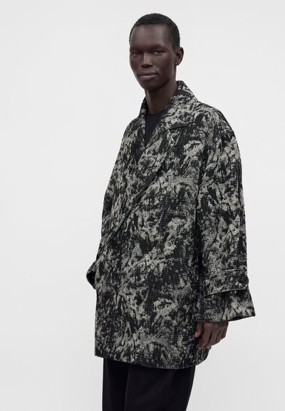 Άνδρας που φοράει ένα μαύρο και γκρι παλτό oversized με διπλό πέτο και ευρύ γιακά, με μαύρο παντελόνι, σε απλό ανοιχτό φόντο.
