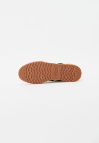 Schwarzer Sneaker mit einer strukturierten Gummisohle, die ein Zickzackmuster aufweist. Die Sohle ist braun und hat runde Griffakzente.