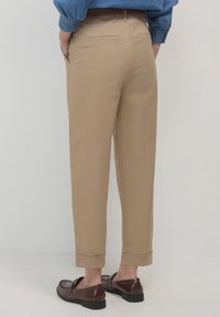 Persona che indossa pantaloni beige con risvolto, mocassini marrone scuro e una camicia di jeans blu, con le mani in tasca, in piedi su uno sfondo neutro.