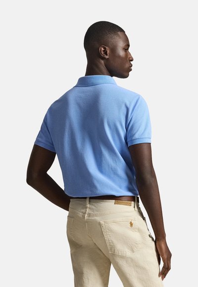 Homme de profil portant un polo bleu clair à manches courtes et un pantalon beige avec une ceinture marron sur un fond uni.