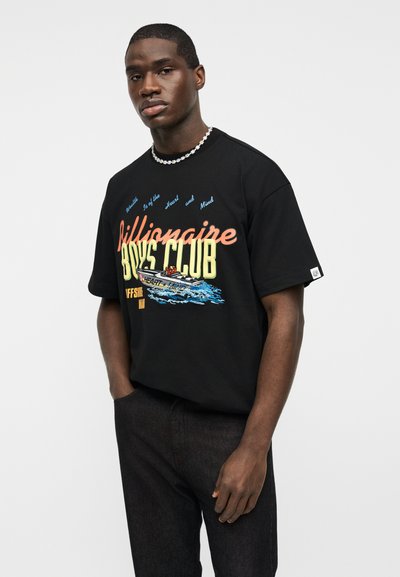 Νέος άνδρας που φοράει μαύρο μπλουζάκι με το λογότυπο Billionaire Boys Club και μια εικονογράφηση πλοίου, συνδυασμένο με σκούρο παντελόνι, μπροστά σε ένα απλό γκρι φόντο.