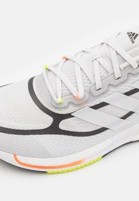 Scarpa sportiva bianca con tomaia in maglia testurizzata, dettagli neri, tre strisce nere e asole per i lacci colorate in giallo, arancione e verde.