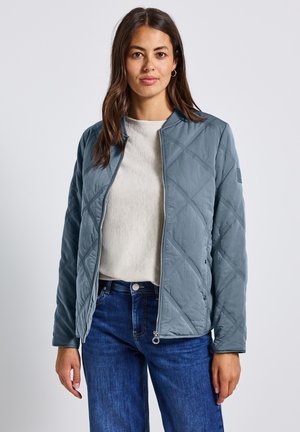 Giacca grigia trapuntata con zip frontale, colletto alto e cuciture a diamante angolate. Indossata sopra un top beige chiaro, abbinata a jeans blu.