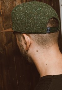 Zöld, textúrált sapka színes foltokkal, barna napellenzővel és állítható pánttal. A háttér egy fából készült felület, ami kontrasztot ad.