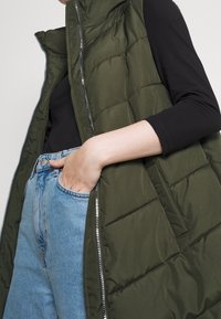 Oliwkowa zielona pikowana kamizelka z wysokim kołnierzem, srebrnym zamkiem błyskawicznym i bocznymi kieszeniami, noszona na czarnej koszuli z długim rękawem i niebieskich dżinsach.