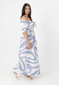 Abito maxi blu e bianco con motivi ondulati, spalle scoperte, vita arricciata e dettaglio a fiocco; abbinato a delle scarpe con tacco aperte.