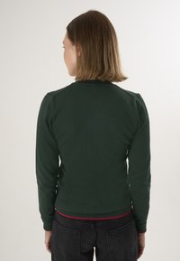 Pull-over vert foncé avec manches longues, col côtelé et accent rouge à l'ourlet. Fabriqué dans un tissu lisse et extensible.