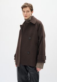 Abrigo de lana marrón con un diseño de doble botonadura, grandes botones y bolsillos laterales. Suéter de punto texturizado debajo, combinado con pantalones de cuero negros.