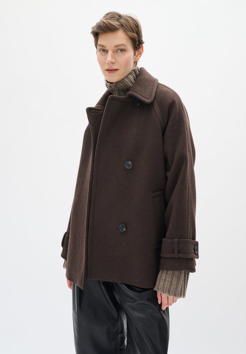 Abrigo de lana marrón con un diseño de doble botonadura, grandes botones y bolsillos laterales. Suéter de punto texturizado debajo, combinado con pantalones de cuero negros.