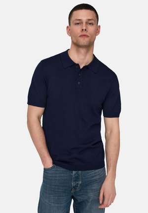 Uomo in piedi con una mano in tasca, indossa una polo a maniche corte blu navy e jeans blu, su uno sfondo chiaro semplice.