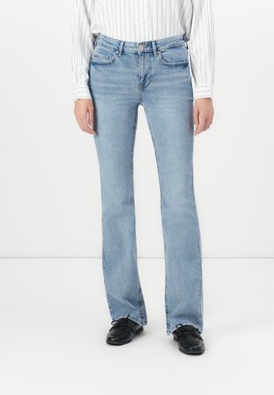 Person som har på seg lyseblå bootcut jeans, svarte snøresko, og en hvit langermet skjorte med tynne vertikale striper.