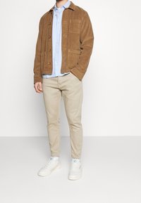 Braune Cordjacke mit Knopfverschluss, hellblaues Hemd mit Kragen, beigefarbene Hose und weiße Sneaker. Das Modell steht vor einem schlichten Hintergrund.