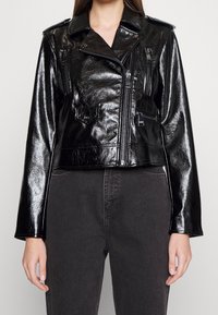 Svart läderbikerjacka med en glänsande finish, utrustad med dragkedjor, bröst- och sidofickor, samt en kortad design.