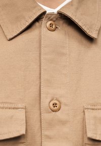 Καφέ πουκάμισο από ντένιμ με κουμπιά στο μπροστινό μέρος, δύο τσέπες στο στήθος και κλασικό γιακά. Το ύφασμα έχει μια διακριτική υφή και ανθεκτική εμφάνιση.