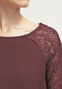 Burgundin värinen paita, jossa on pitsiyksityiskohtia hihoissa, esittelee monimutkaisia kukkakuormia ja pehmeää, sileää kangasta vartalossa.