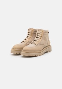 Botas de tornozelo em camurça bege com colarinho acolchoado, ilhós dourados, atacadores planos e uma sola espessa e robusta com padrões pronunciados.