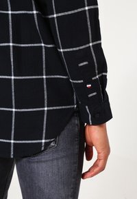 Maglietta nera a maniche lunghe con motivo a finestra bianca indossata con jeans grigio scuro, che mostra il polsino con un piccolo bottone rotondo rosso, bianco e blu.