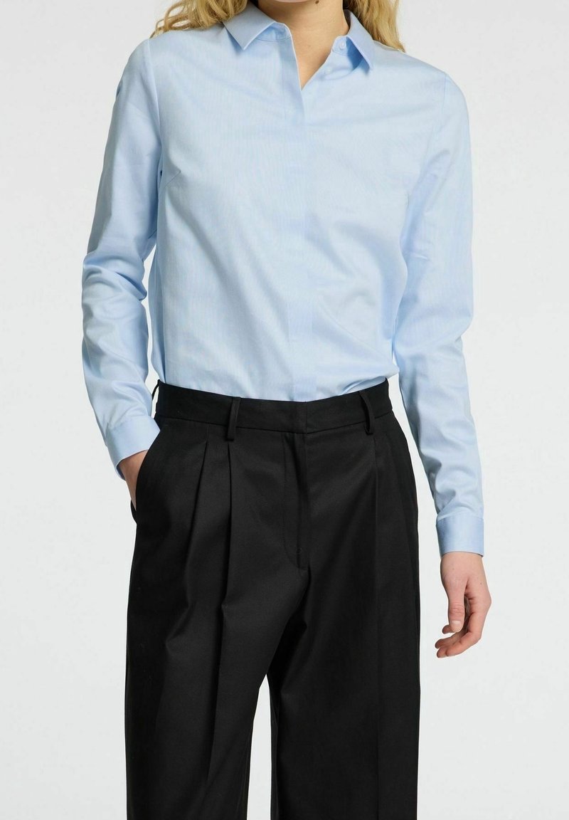 Camicia azzurra a maniche lunghe con colletto, abbinata a pantaloni neri plissettati. La camicia presenta un tessuto liscio e una vestibilità aderente.