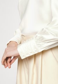 Primo piano di mani che regolano il polsino di una blusa di satin bianco con bottoni rivestiti di tessuto, abbinata a pantaloni cream a vita alta.