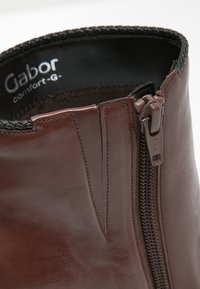 Ruskea nahkainen nilkkabootsi, jossa on sivuvetoketju ja teksturoitu reunus. Ominaisuuksiin kuuluu sileä pinta ja ommeltu yksityiskohta yläreunassa.