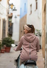 Felpa rosa chiaro con cappuccio e dettagli con zip, abbinata a un motorino blu vintage, in una strada stretta con vegetazione e edifici.