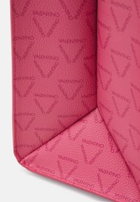 Růžová texturovaná kůže s opakujícím se vzorem loga Valentino, který vytváří tvary srdcí, zobrazená na přehnutém rohu produktu.