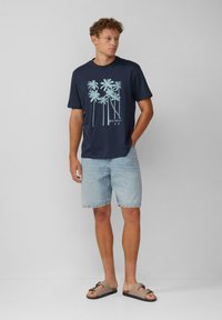 Marineblaue Baumwoll-T-Shirt mit einem hellblauen Palmenmotiv, kombiniert mit hellen Denim-Shorts und braunen Sandalen mit einer Schleifenakzent.