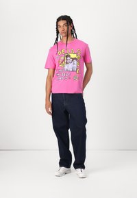 Pink cotton t-shirt featuring a graphic print with text, flowers, and a photo design. Worn with dark jeans and white sneakers.