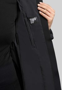 Zwarte pufferjas met een zachte afwerking, met een stoffen label aan de binnenkant. Het ontwerp heeft een gestroomlijnde silhouet en verborgen zakken.