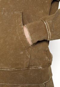 Sudadera marrón con una superficie texturizada, que cuenta con un bolsillo frontal, puños acanalados y dobladillo. La tela presenta un aspecto descolorido y gastado.