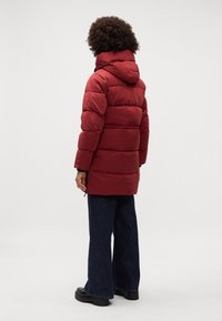 Rød polstret jakke med hætte, der har et quiltet design og sidelommer. Parret med bukser i brede ben i mørk denim og kraftige sorte sko.