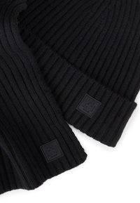 Czarny, prążkowany, dziany zestaw szalika i czapki z małymi gumowymi logo marki w kształcie kwadratu przyszytymi blisko krawędzi na białym tle.