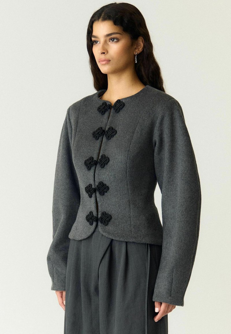 Femme portant une veste gris foncé cintrée avec des fermoirs décoratifs noirs et un pantalon large assorti, debout devant un fond uni.