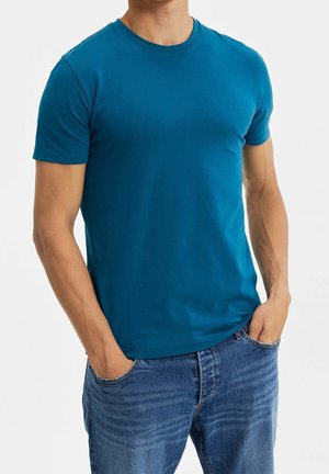 Vihreään vivahtava puuvillainen t-paita, jossa pyöreä pääntie ja lyhyet hihat, yhdistettynä sinisiin farkkuihin. Kangas näyttää sileältä ja koristelemattomalta.