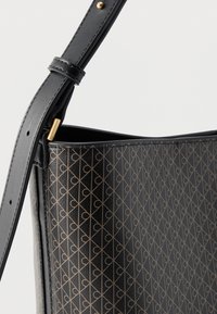 Μαύρη δερμάτινη τσάντα χειρός με ρυθμιζόμενο λουρί, που διαθέτει επαναλαμβανόμενο γεωμετρικό μοτίβο σε ταμπά χρώμα και αγκράφα από μέταλλο σε χρυσή απόχρωση.