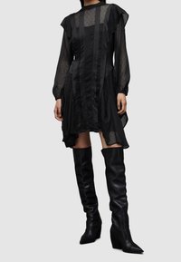 Femme portant une robe longue noire transparente à manches longues avec un motif à pois, assortie à des bottes en cuir noires montant jusqu'aux genoux.
