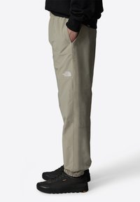 Beige vattentäta byxor med elastiska ärmslut, utrustade med sidofickor och en diskret logotyp på låret. Bärs med svarta utomhusskor.