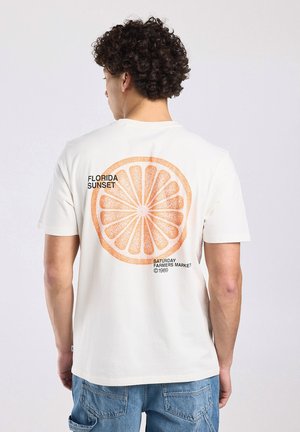 Man with curly hair wearing white T-shirt showing large orange slice graphic and text "Florida Sunset" on back.