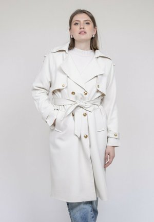 Femme portant un trench-coat blanc ceinturé avec des boutons dorés, par-dessus un pull blanc et un jean bleu clair, debout devant un fond uni.