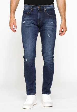 Uomo con jeans blu slim fit consumati e sneaker bianche, in piedi davanti a uno sfondo bianco semplice.