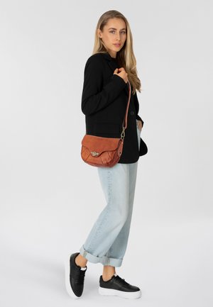 Borsa a tracolla in pelle marrone con patta rotonda, dotata di una chiusura metallica, posizionata su un modello che indossa un blazer nero e jeans azzurri.