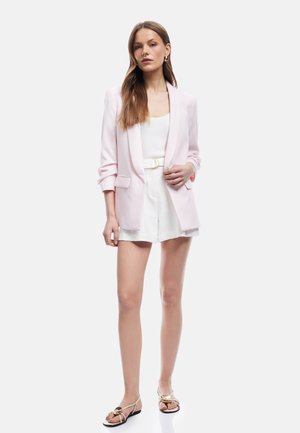 Femme portant un blazer rose clair sur un top blanc sans manches et un short blanc taille haute avec ceinture, associé à des sandales blanches.