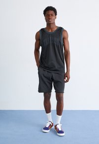 Svart ermeløs trenings tank topp med blå kant og "ELLESSE" logo, kombinert med grå shorts og mønstrede joggesko, stående på en blå bakgrunn.