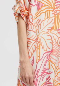 Weiblicher Arm, der neben einem Stoff mit orangefarbenen und rosa tropischen Blattmustern hängt, mit hochgekrempeltem Ärmel.